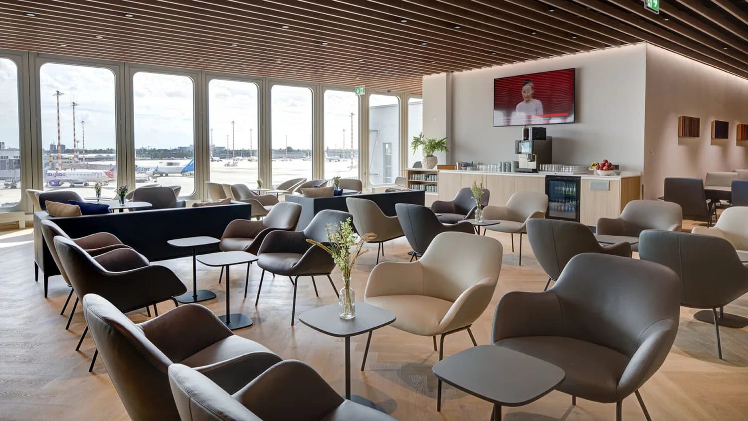 Lounge im Flughafen mit Sitzgruppen, Tischen, Panoramafenstern zum Vorfeld, Flugzeuge im Hintergrund und Selbstbedienungsbereich mit TV und Getränken
