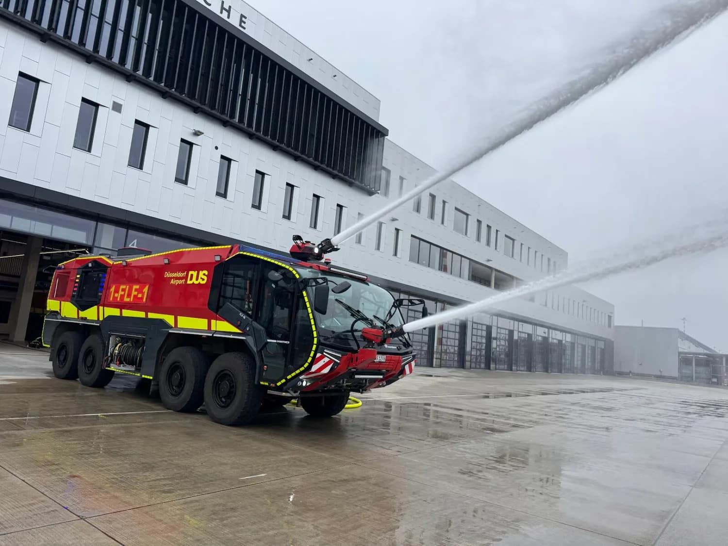 Rotes Airport-Feuerwehrfahrzeug DUS am Düsseldorfer Flughafen spritzt Wasser aus Dach- und Frontmonitor vor Hangargebäude