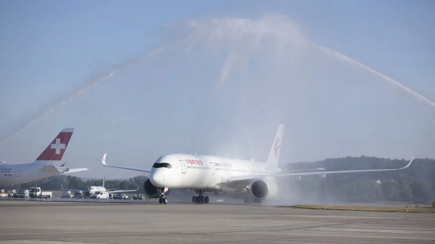China Eastern Passagierflugzeug bei Erstflug empfängt traditionelle Wasserfontänen-Taufe am Flughafen, Seitenansicht auf Rollfeld mit Flughafenfahrzeugen und Schweizer Flugzeugheck im Hintergrund