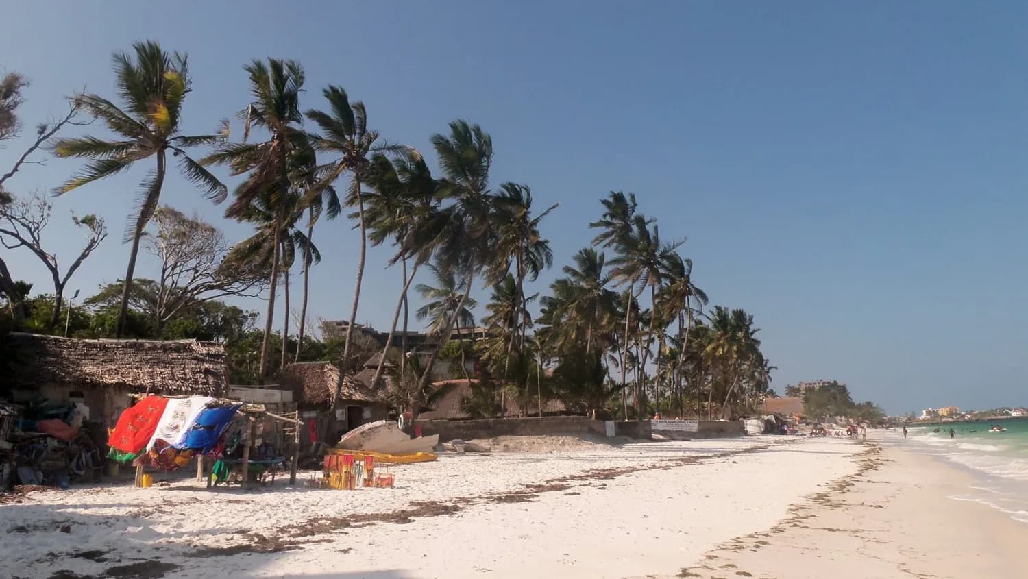 Strand von Mombasa mit weißem Sand, Palmenreihe, Strohhütten und bunten Verkaufsständen am Ufer