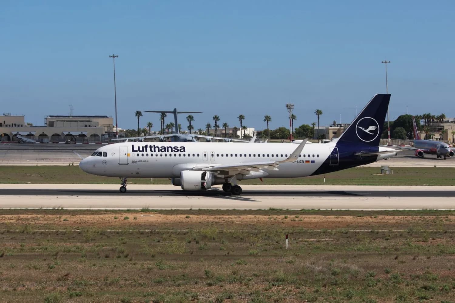 Lufthansa Airbus A320 auf Rollbahn am Flughafen, Seitenansicht mit Triebwerken und dunkelblauer Heckflosse vor Terminal und Taxiway