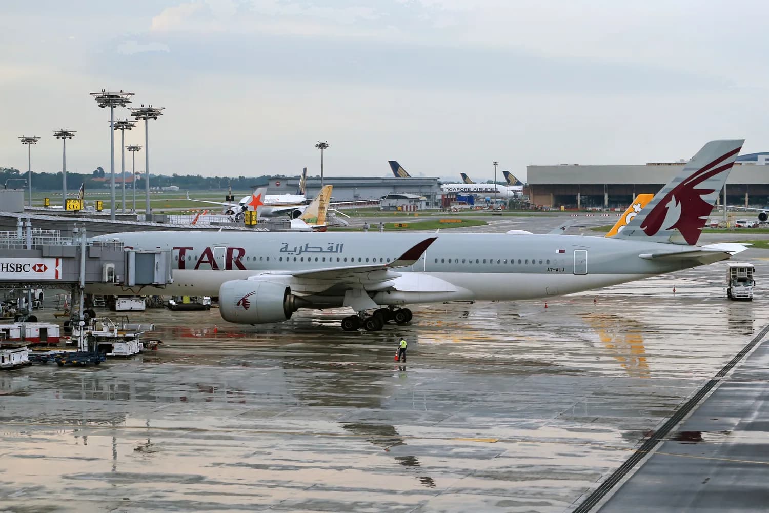 Qatar Airways Passagierflugzeug am Gate eines Flughafenterminals bei regennasser Apronfläche, Bodenpersonal und weitere Flugzeuge im Hintergrund