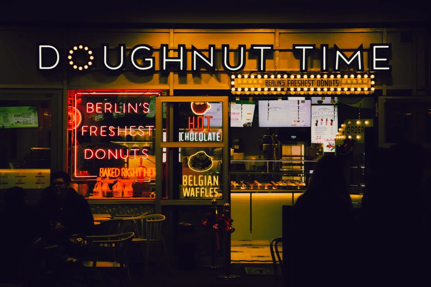 Dunkel beleuchtete Donut-Bude in Berlin mit Neon-Schildern „Doughnut Time“, „Berlin’s freshest donuts“, „Hot Chocolate“ und „Belgian Waffles“ sowie Tresen und Sitzbereich