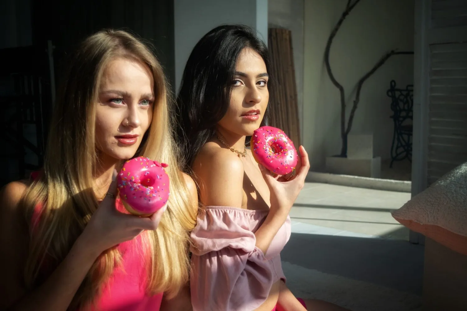Zwei junge Frauen sitzen nebeneinander und halten pink glasierte Donuts mit Streuseln, modisches Lifestyle-Foto, Innenraum mit Sonnenlicht in Berlin