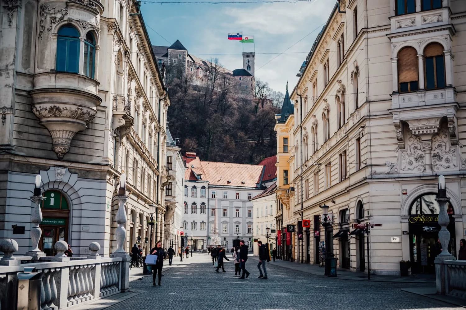 Breite Fußgängerstraße in der Altstadt von Ljubljana mit historischer Architektur, Menschen, Brunnen und Burg auf dem Hügel im Hintergrund