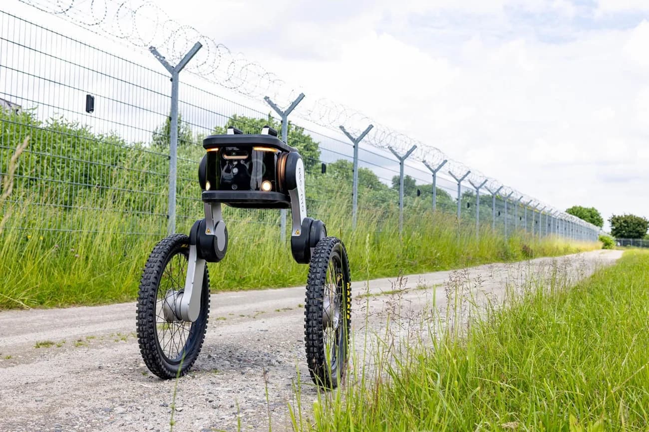 Sicherheitsroboter auf zwei geländetauglichen Rädern patrouilliert neben Hochsicherheitszaun mit Stacheldraht auf Schotterweg in ländlicher Umgebung