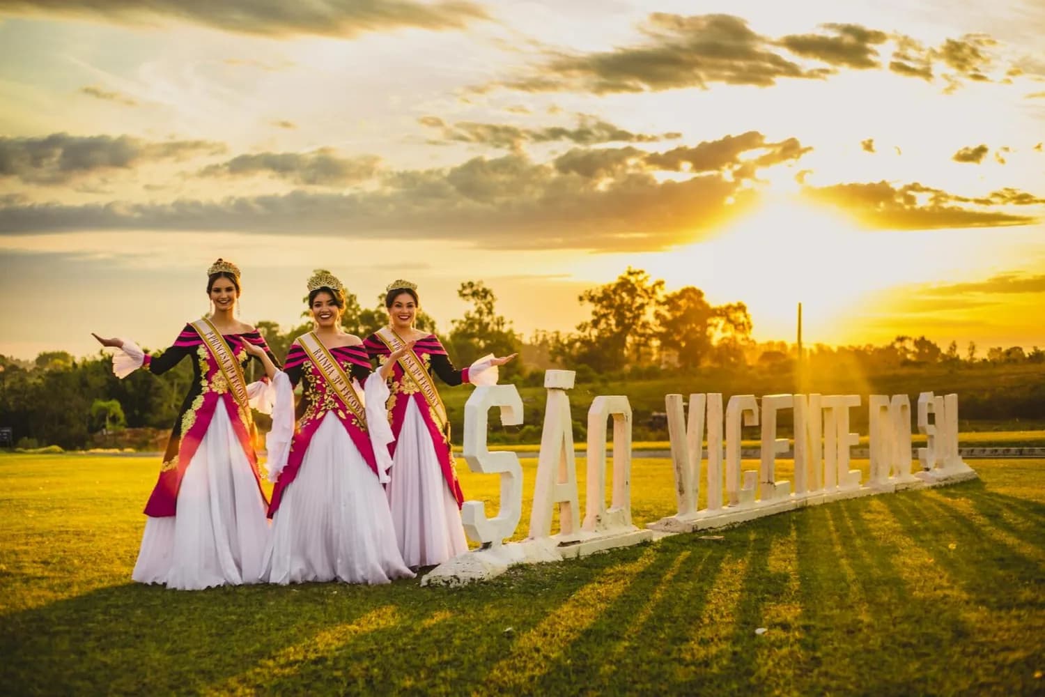 Drei Schönheitsköniginnen in traditionellen rosa-weißen Kleidern und goldenen Kronen posieren vor dem beleuchteten Schriftzug São Vicente bei Sonnenuntergang auf Kap Verde.