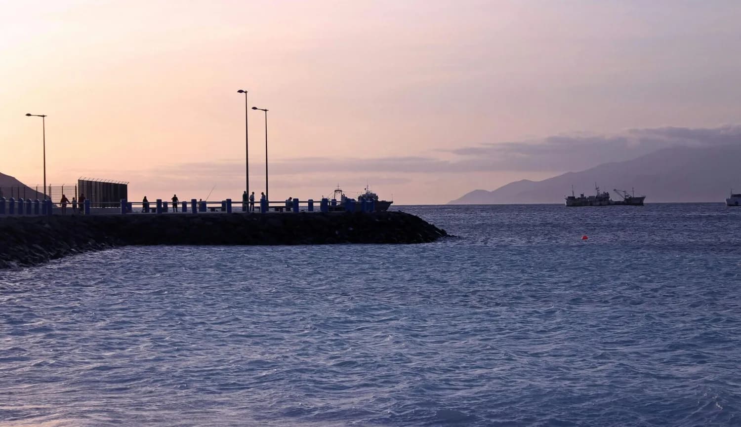 Mole mit Fußgängern und Anglern am Hafen von Mindelo, Kapverden bei ruhigem Meer und Sonnenuntergang, Fischerboote in der Bucht vor bergiger Küste