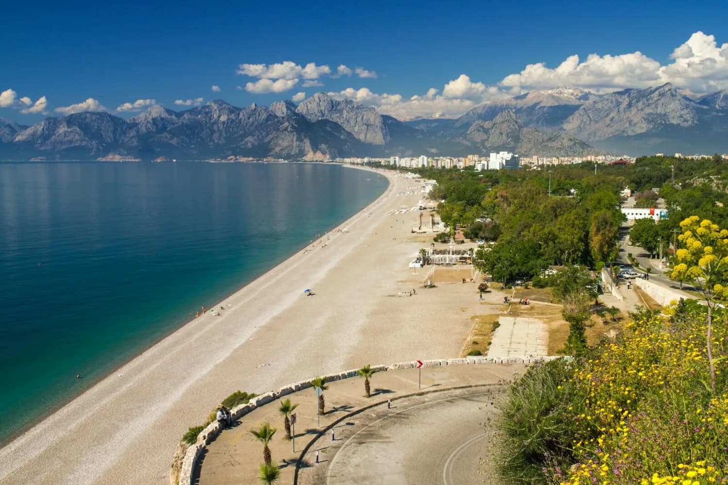 Antalya Küstenpanorama mit langem Kiesstrand, türkisblauem Meer, Strandliegen, Pinien und Stadt am Fuße schroffer Taurusgebirge – Urlaubskulisse am Mittelmeer