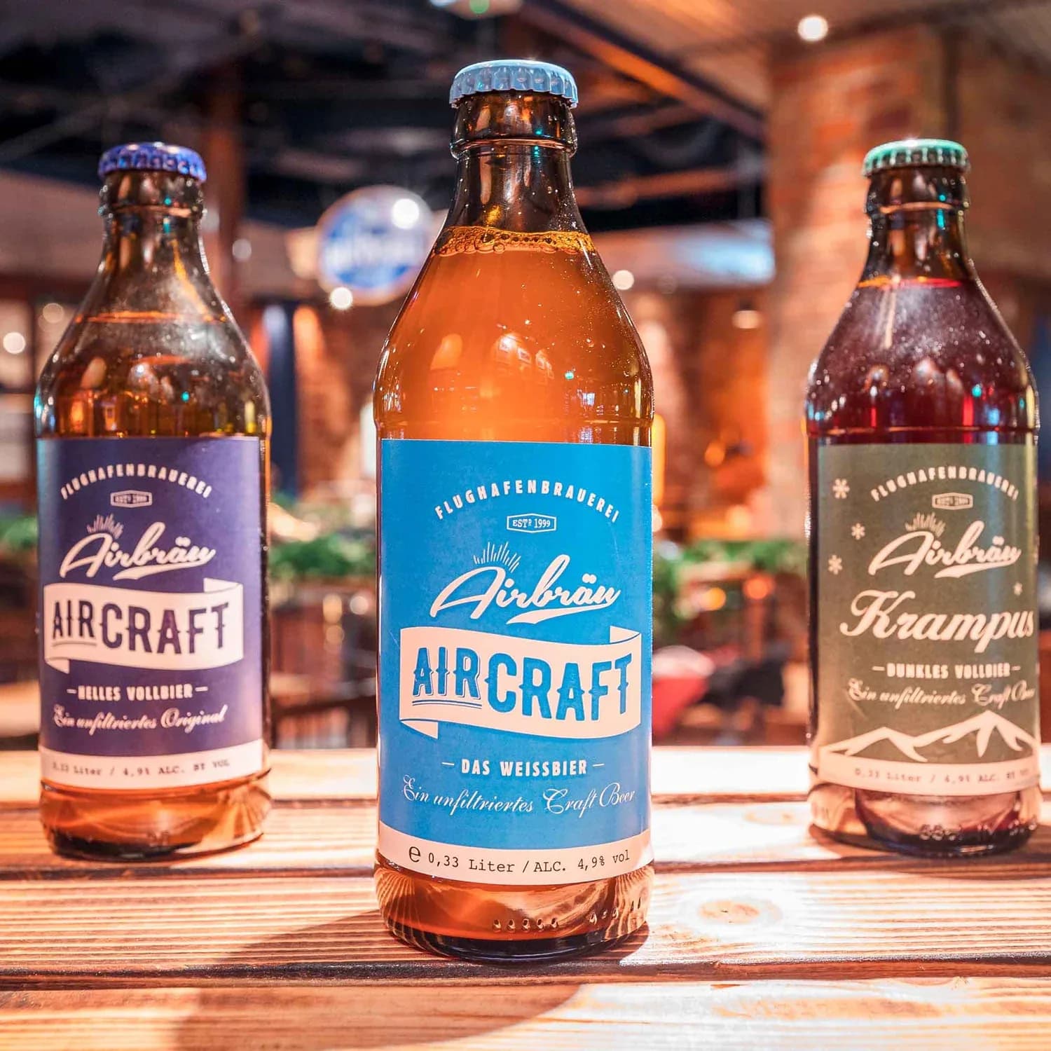 Drei Airbräu Bierflaschen im Vordergrund, mittlere mit blauem Aircraft-Label, links helles Aircraft, rechts dunkles Krampus, auf Holztisch in Brauerei-Umgebung