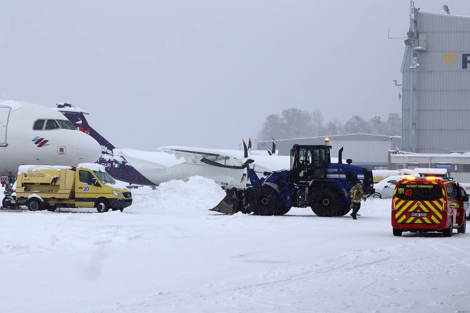 Radlader und Flughafenfahrzeuge räumen Schnee am verschneiten Rollfeld des Flughafens Nürnberg neben geparkten Propellerflugzeugen und Einsatzwagen