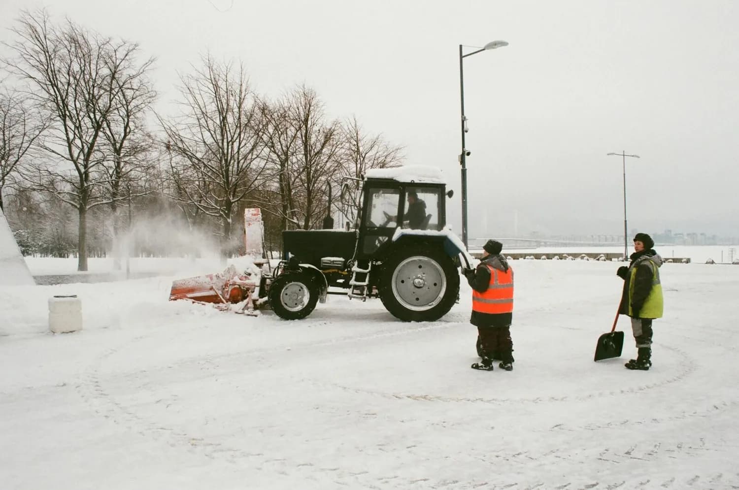 Winterlicher Traktor mit Schneepflug räumt verschneite Promenade, zwei Winterdienstmitarbeiter mit Warnwesten und Schaufel daneben