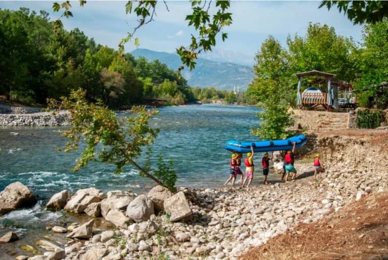 Gruppe beim Rafting trägt Schlauchboot am Ufer des Köprüçay im Köprülü-Kanyon bei Antalya, felsige Flusslandschaft mit Bäumen und Gebirgsblick
