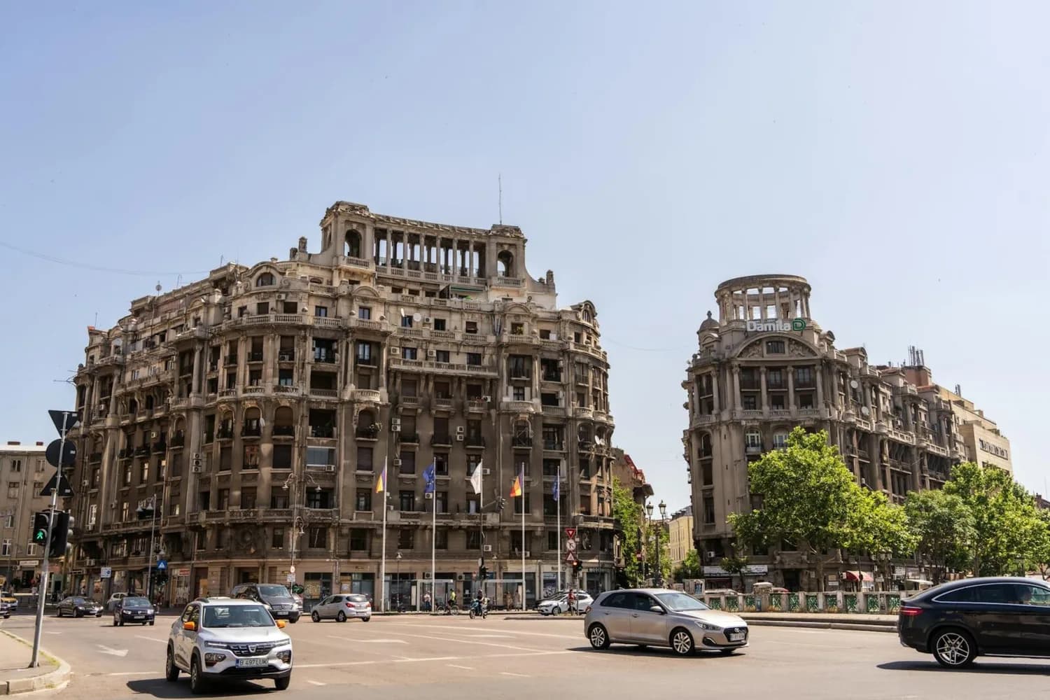 Breite Aufnahme historischer Jugendstil- und Neoklassik-Gebäude an einem belebten Straßenkreuz in Bukarest mit Autos, Bäumen und Fahnen vorm klaren Himmel