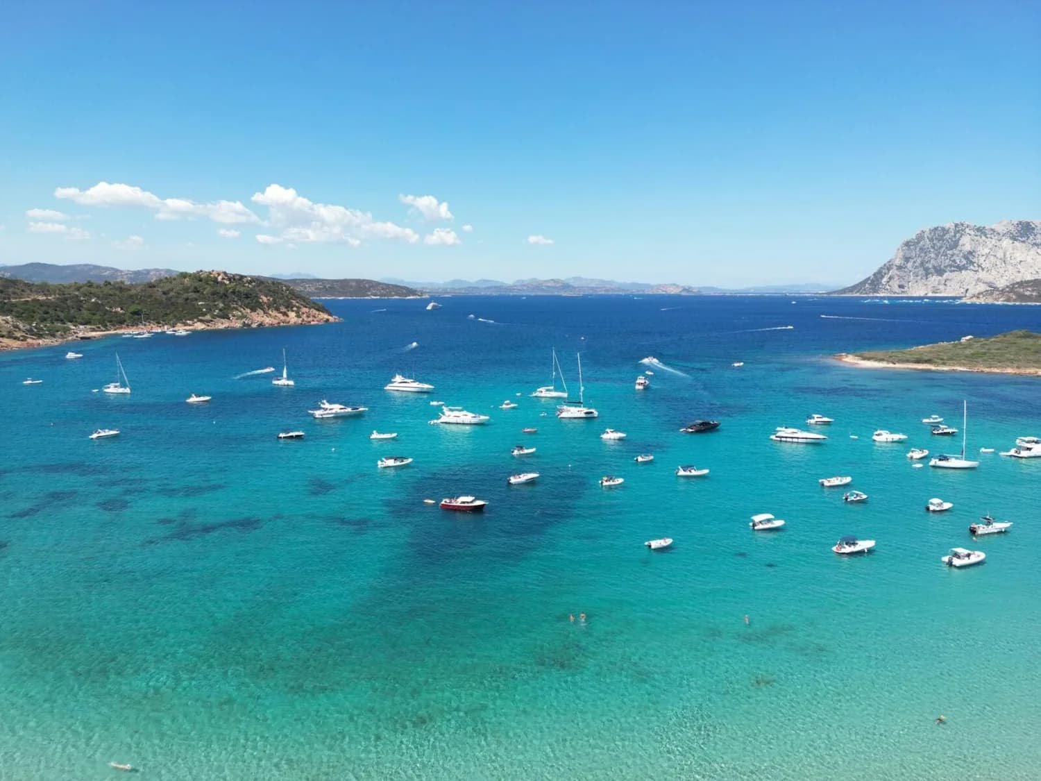 Bucht an der Smaragdküste Sardiniens mit türkisblauem Wasser und vielen Ankerliegern und Segelbooten vor felsiger Küste und klarem Himmel