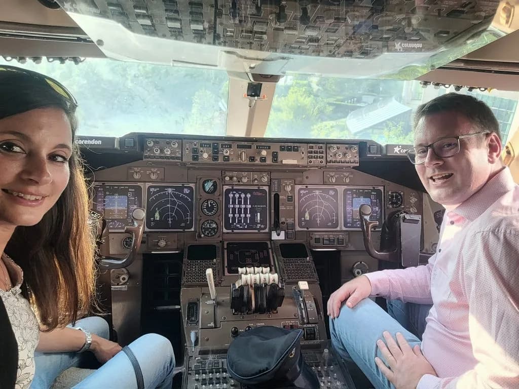 Zwei lächelnde Personen im Cockpit eines Verkehrsflugzeugs vor Instrumententafel und Schubhebeln, Pilotenkappe auf dem Boden.