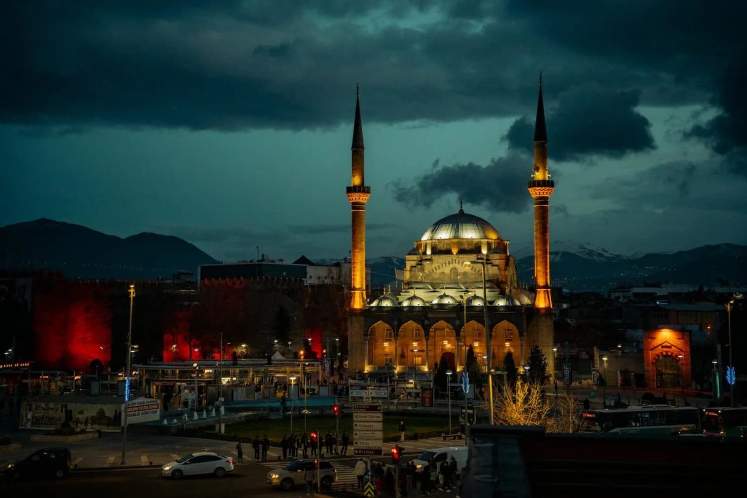Beleuchtete Selimiye-Moschee mit zwei Minaretten bei Dämmerung in Kayseri, Türkei, Stadtzentrum mit Verkehr und Bergen im Hintergrund