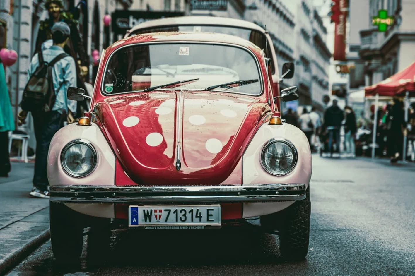 Roter und rosa VW-Käfer mit weißen Punkten auf einer belebten Wiener Straße, Frontansicht mit Wiener Kennzeichen und Fußgängerzone im Hintergrund