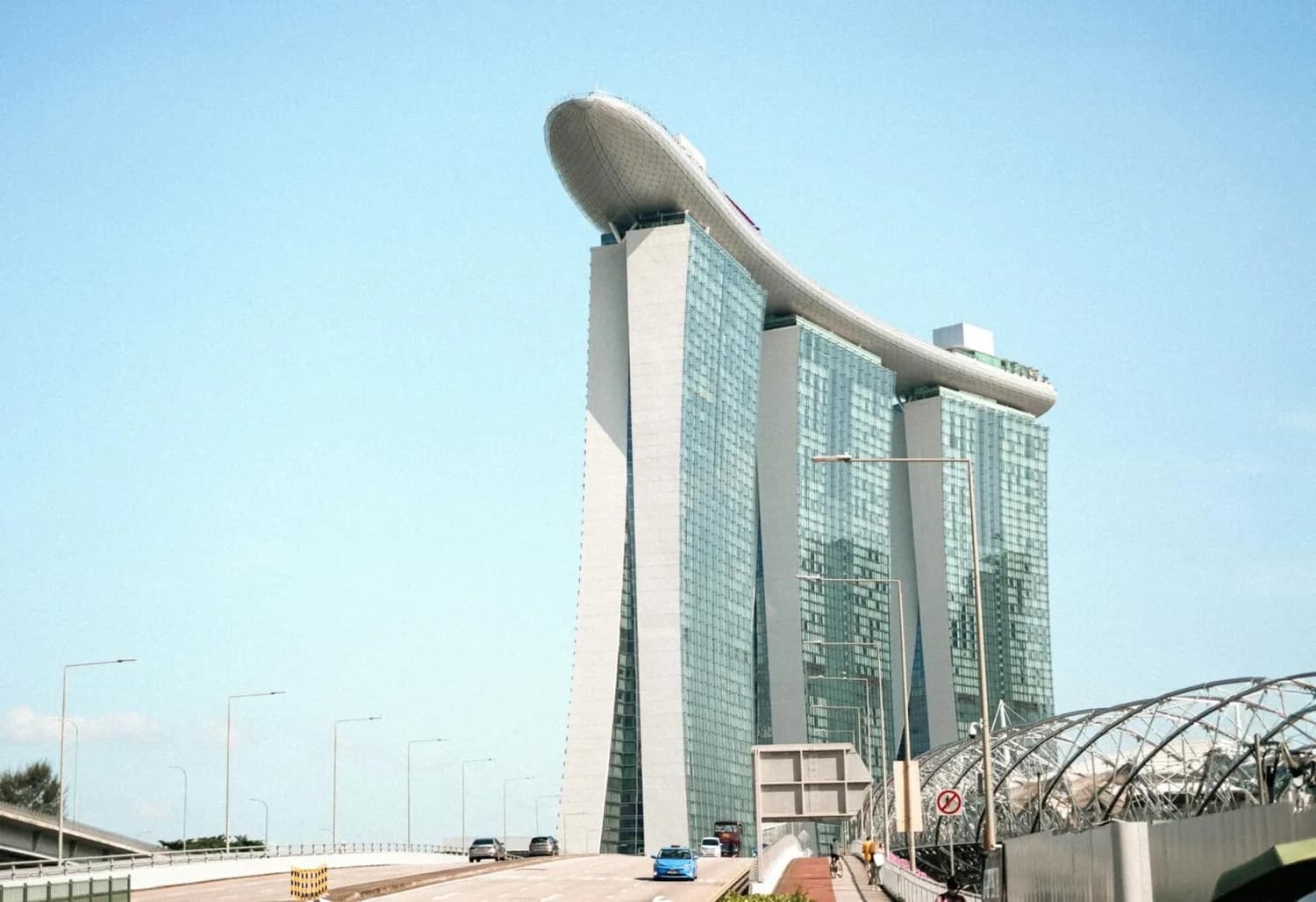 Marina Bay Sands Hotel in Singapur mit markanter SkyPark-Dachterrasse, Glasfassade, darunter Fahrspur und Fußweg im Vordergrund