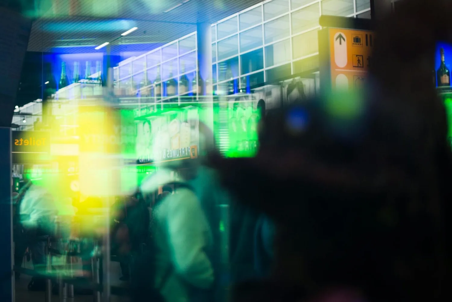 verspiegelte Aufnahme von Flughafenhalle mit Passagieren, Sicherheitskontrolle und Leitschild, bunte Neon- und Reflexionseffekte