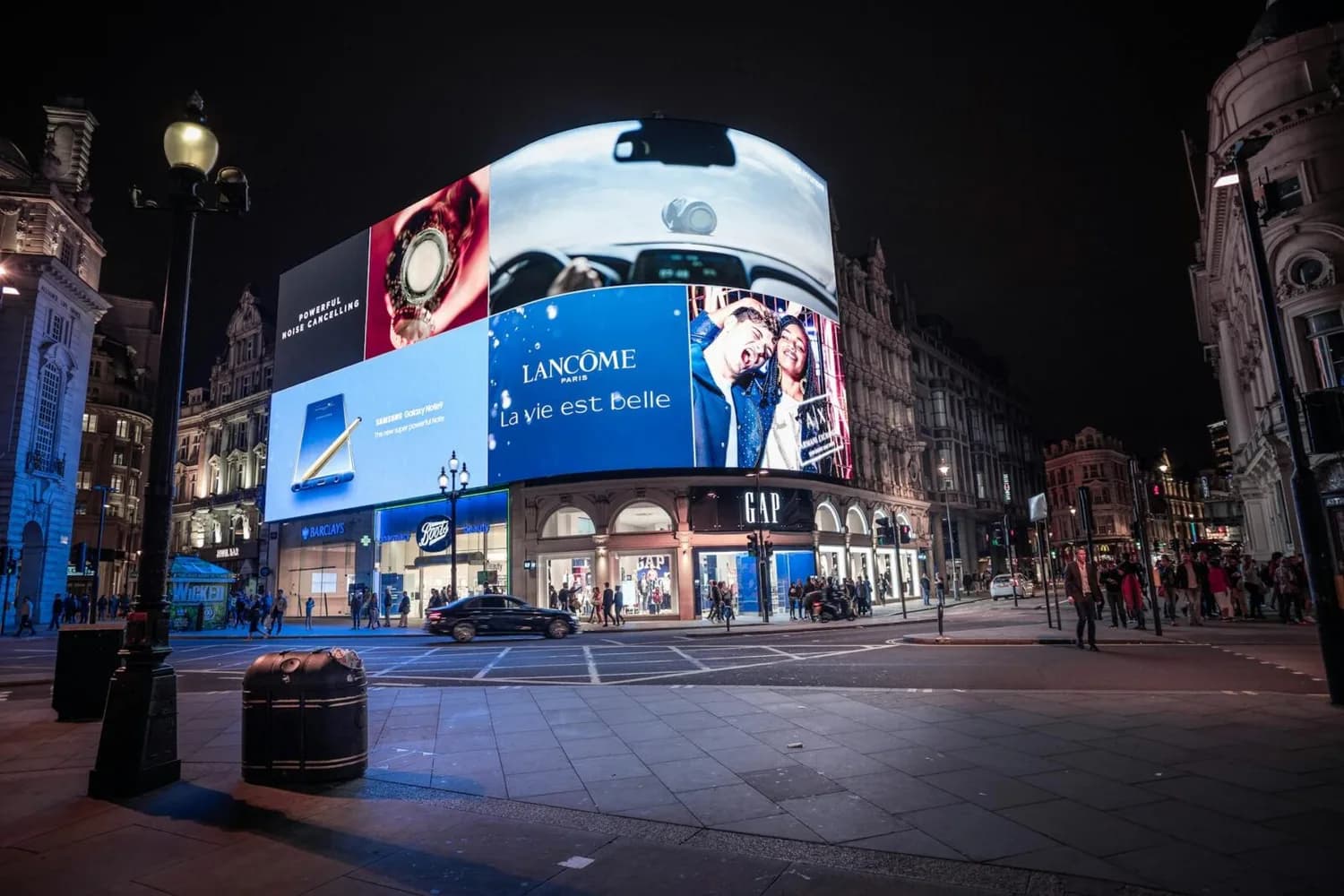 Nächtliche Stadtkreuzung mit großer gebogener DOOH-Leinwand über GAP-Shop, beleuchtete Werbeflächen (Lancôme, Samsung), Fußgänger und Verkehr in einer europäischen Innenstadt
