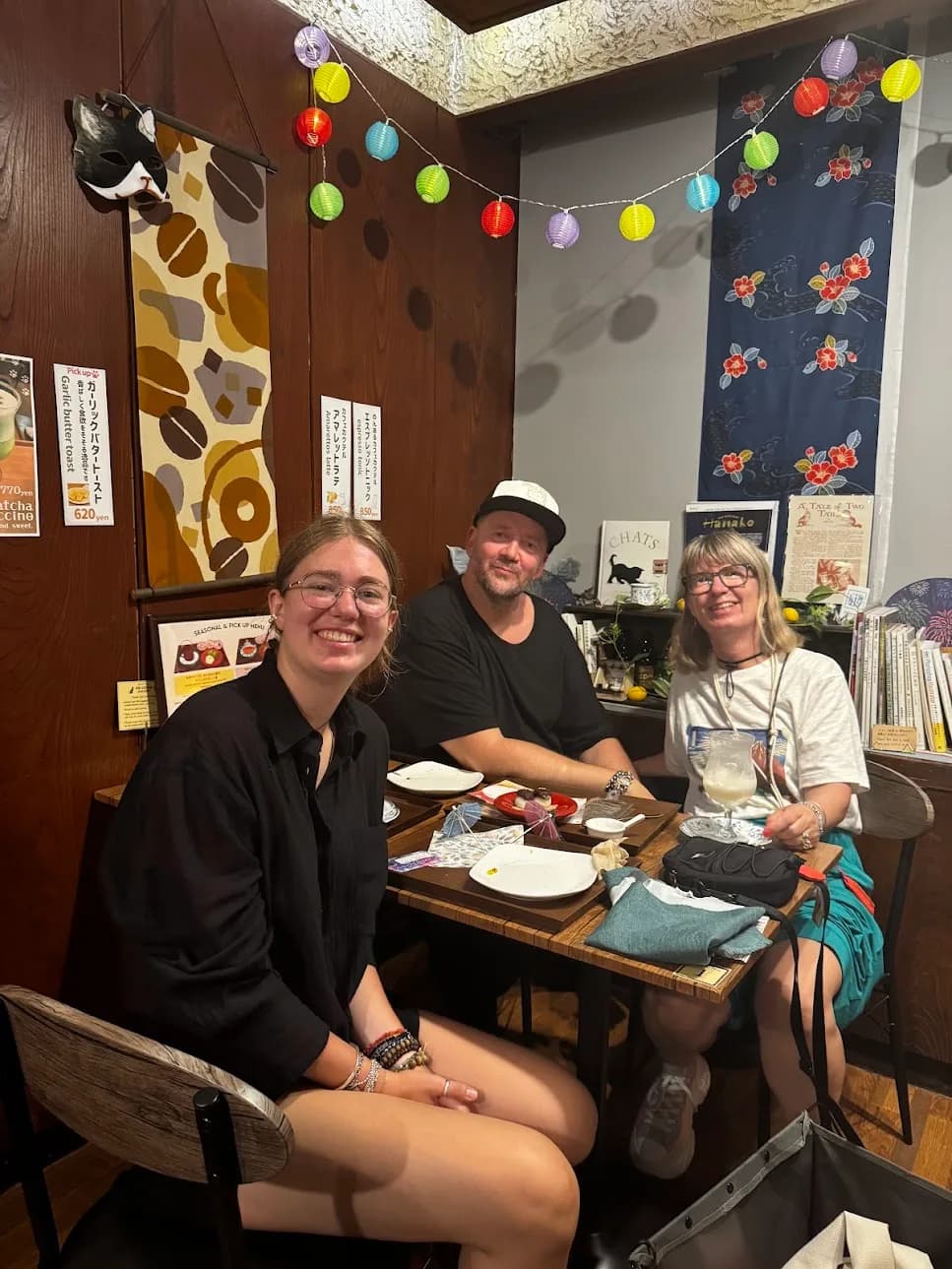 Drei Personen lächelnd an einem Tisch in gemütlichem japanischen Café mit Laternenkette, Wandbehang mit Blumenmotiv, Speisekarten und Geschirr auf dem Tisch