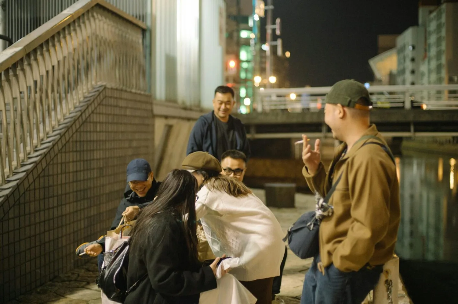 Gruppe japanischer Freunde lacht und teilt Snacks an einer nächtlichen Uferpromenade in Japan, entspannte Omotenashi-Gastfreundschaftsszene