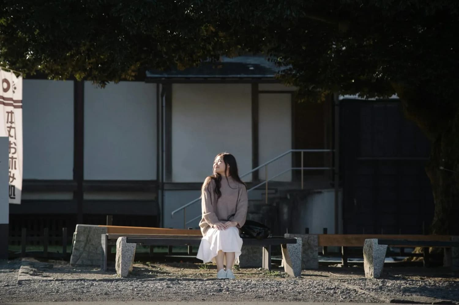 Junge Frau sitzt auf Holzbank unter Baum vor traditionellem japanischen Gebäude und blickt entspannt zur Sonne, Omotenashi-Szene