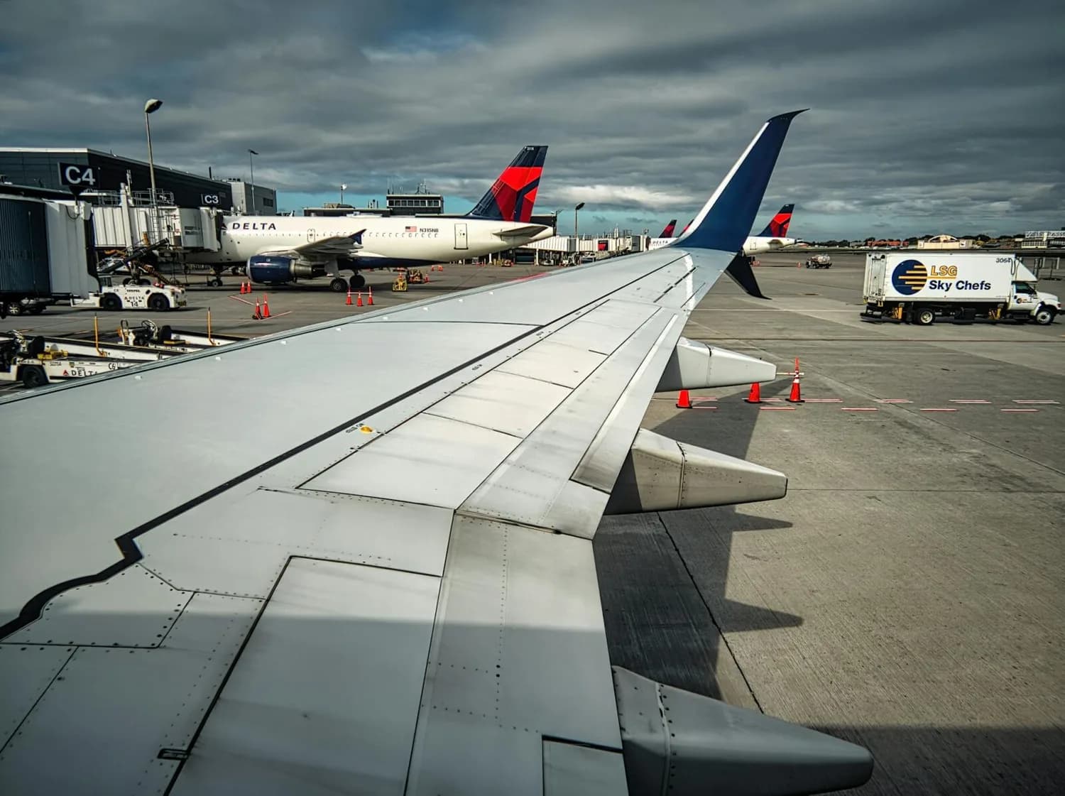 Blick aus Fenster auf Flugzeugtragfläche und Winglet am Flughafen mit geparkten Delta-Maschinen und Catering-LKW auf dem Vorfeld