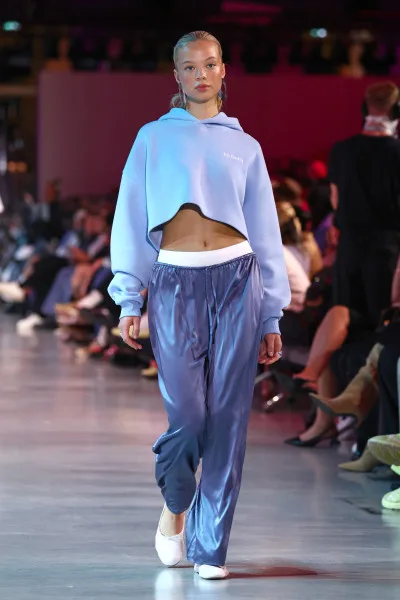 Model läuft auf Modenschau: junge Frau trägt hellblauen, kurz geschnittenen Hoodie, weiße Sporthoseinlage und glänzende blaue Satin-Jogginghose, Sneaker, Laufstegszene mit Publikum. (c) Eurowings