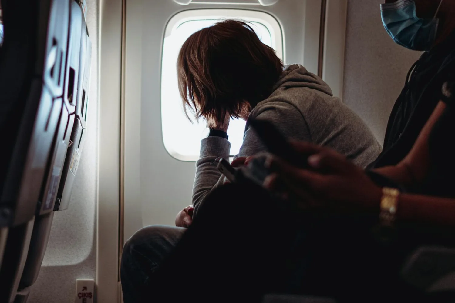 Passagier mit Kapuzenpullover lehnt nervös am Fensterplatz im Flugzeug, Hand ans Gesicht, neben ihm weitere Sitznachbarn und Smartphone sichtbar