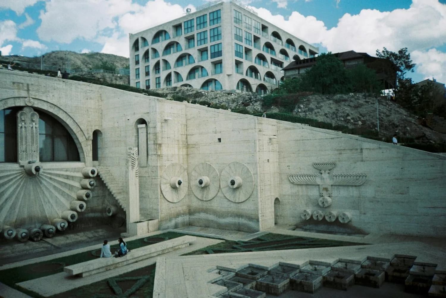 Brunnenplatz mit skulpturalen Reliefs und runden Wasserdüsen, moderner mehrstöckiger Wohnbau im Hintergrund, Eriwan Armenien