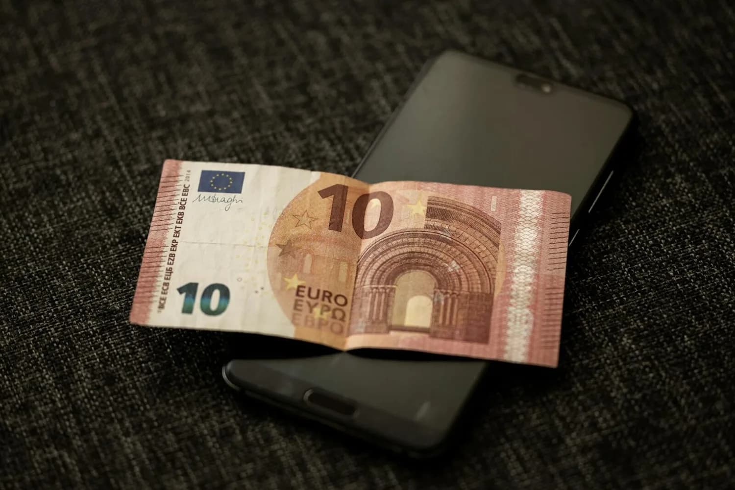 10-Euro-Schein liegend auf schwarzem Smartphone auf dunklem Stoffbezug