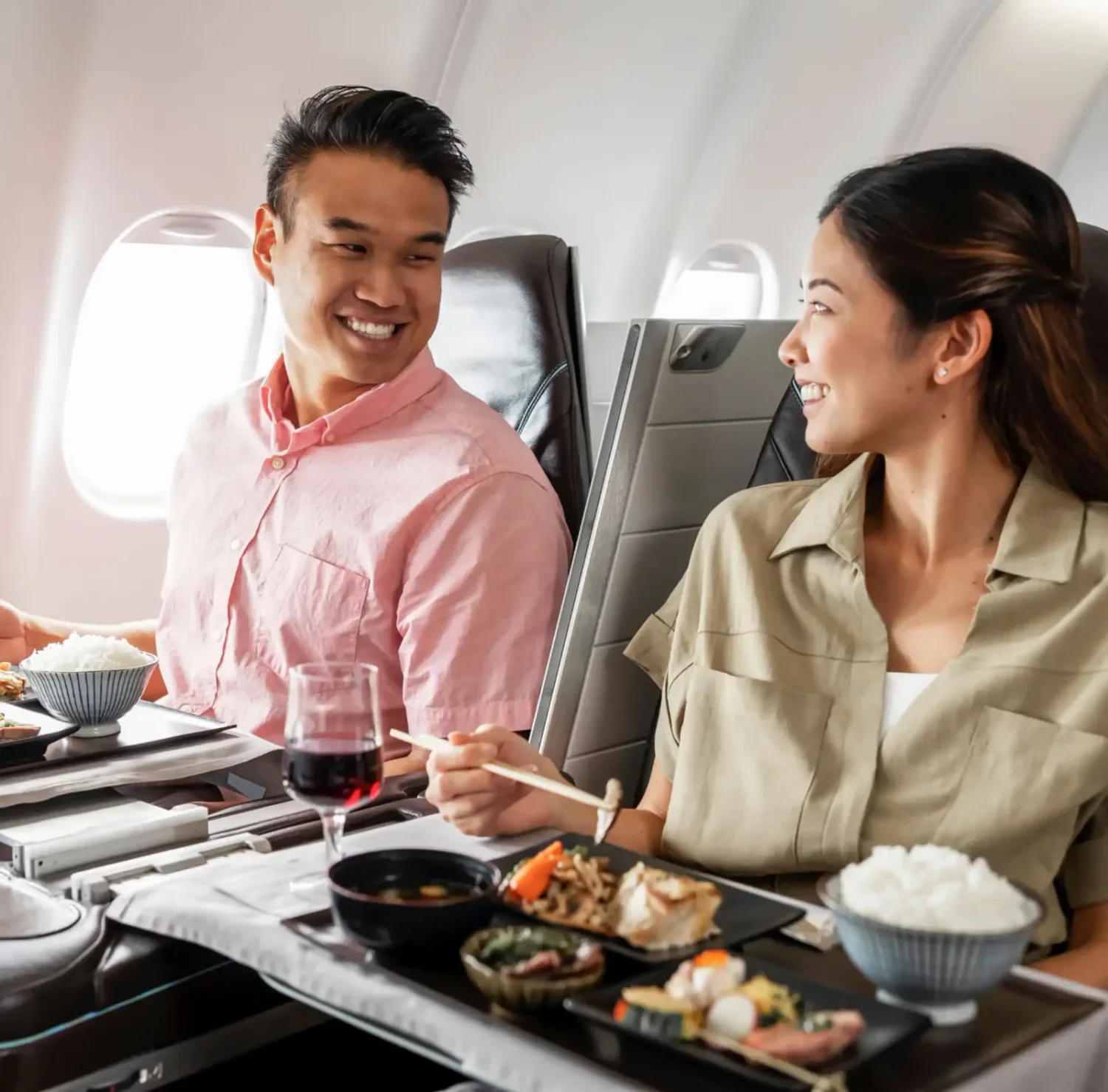 Zwei Passagiere in Flugzeug-Business-Class lächeln sich an beim Servieren eines mehrgängigen Menüs mit Reis, Vorspeisen und Getränken (Chef series2x)