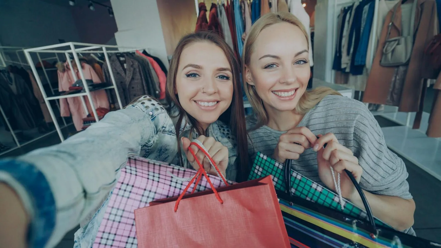 Zwei lächelnde Frauen beim Einkaufen im Airport-Shop mit mehreren Einkaufstüten und Kleidungständern im Hintergrund