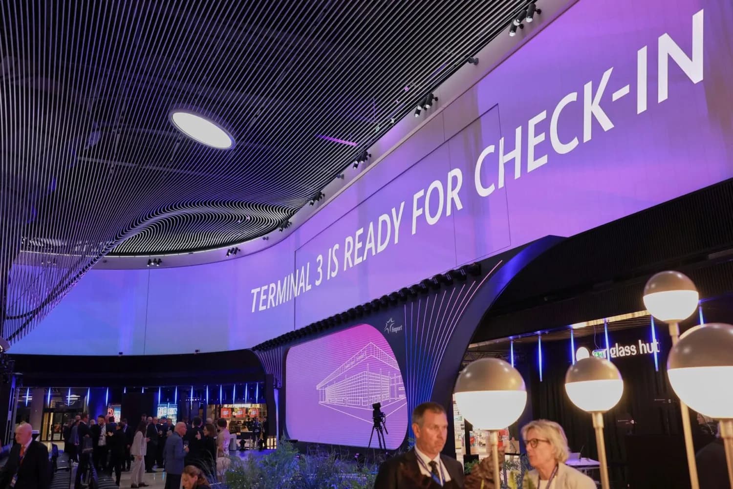 Innenansicht eines Flughafenterminals mit großem violettem digitalen Display „Terminal 3 is ready for check-in“, geschwungener Deckeninstallation und Reisenden am Check-in-Bereich
