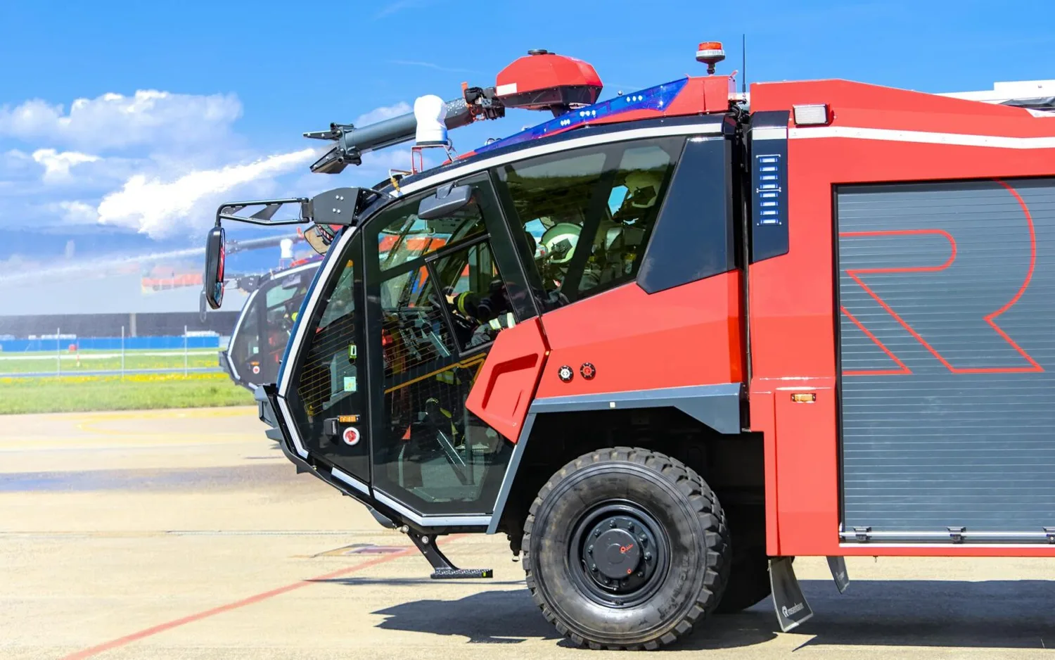 Rotes Flughafenlöschfahrzeug im Einsatz auf Rollfeld mit sichtbarem Fahrerhaus, seitlicher Geräteraum und Löschmonitor, Flughafen und Himmel im Hintergrund