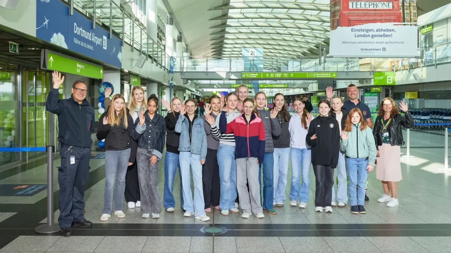 Gruppenbild von Mädchen und Begleitpersonen beim Girls'Day im Flughafen-Terminal, alle stehen in Reihe und winken in die Kamera