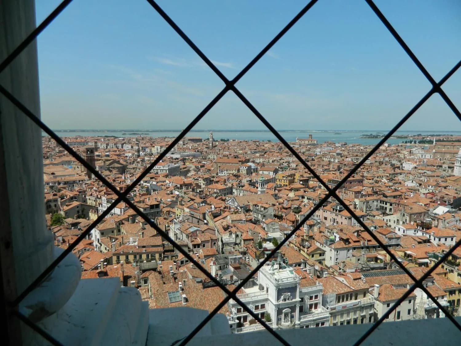 Vogelperspektive auf Venedig mit roten Dachziegeln, Kanälen und Lagune hinter Gittern eines Aussichtsturms
