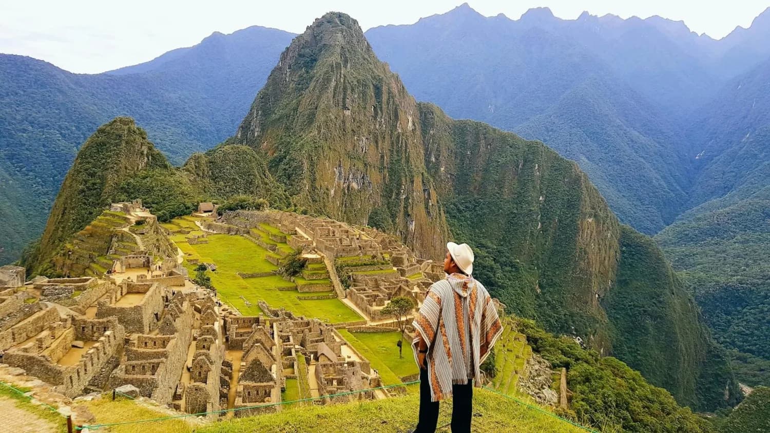 Mann in Poncho und Hut blickt auf die Inka-Ruinen von Machu Picchu mit Terrassen und Bergkulisse