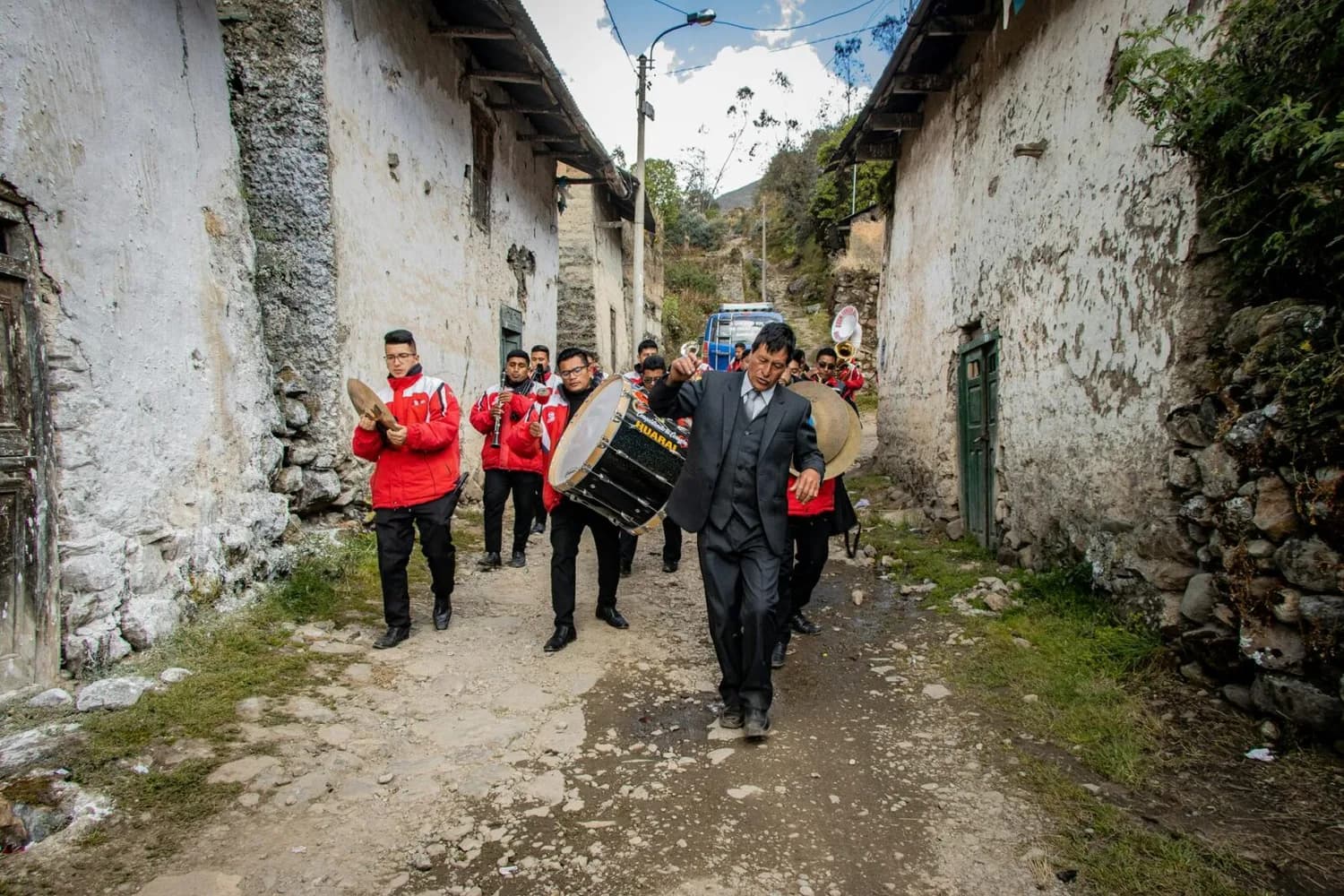 Peruanische Blaskapelle in roter Kleidung und schwarzem Anführer in Anzug beim Marsch durch enges Dorf mit weiß getünchten Häusern und unbefestigtem Weg