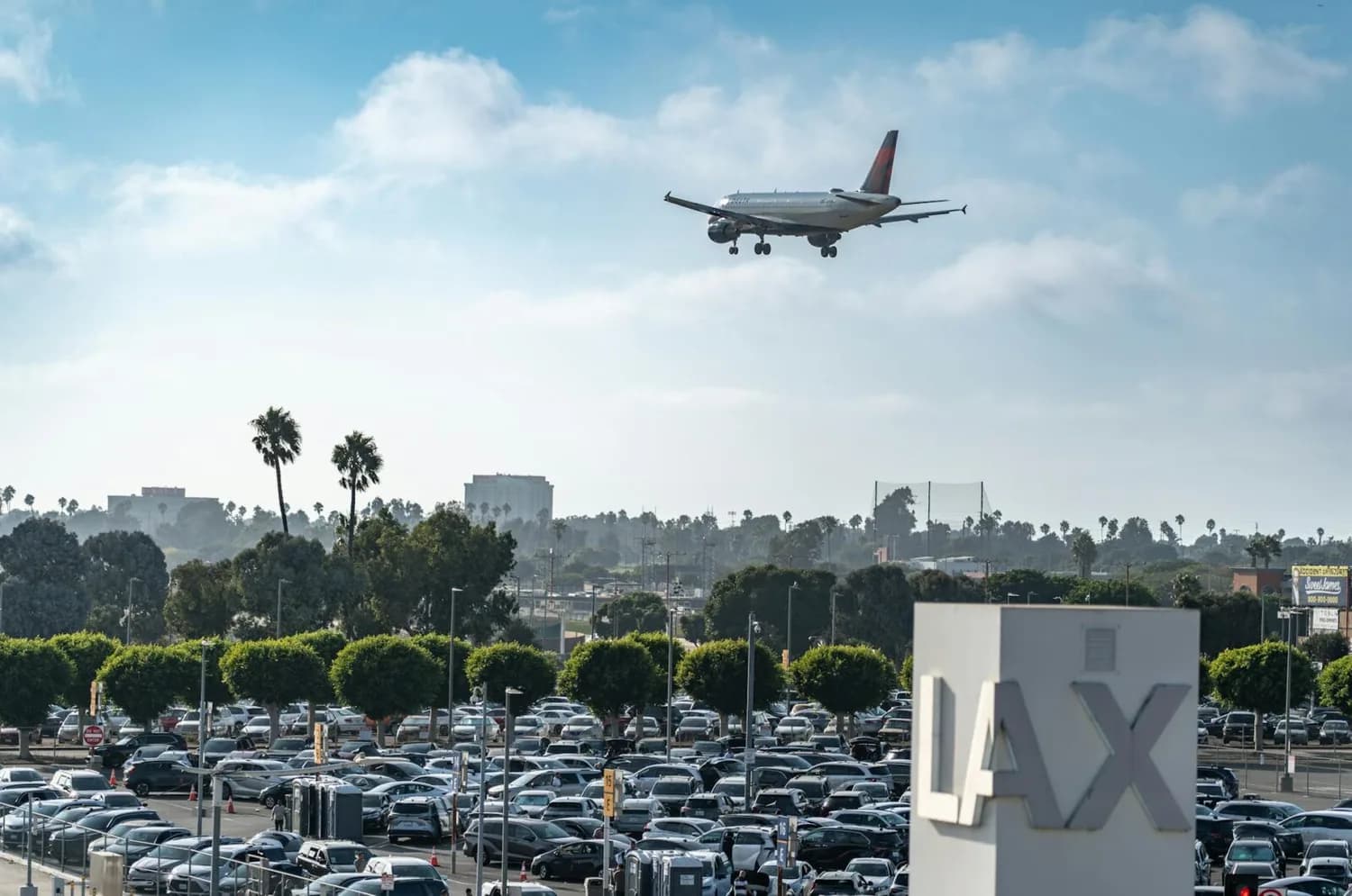 Flugzeug im Landeanflug über vollen Parkplatz am Flughafen Los Angeles LAX mit Palmen und Stadtsilhouette im Hintergrund