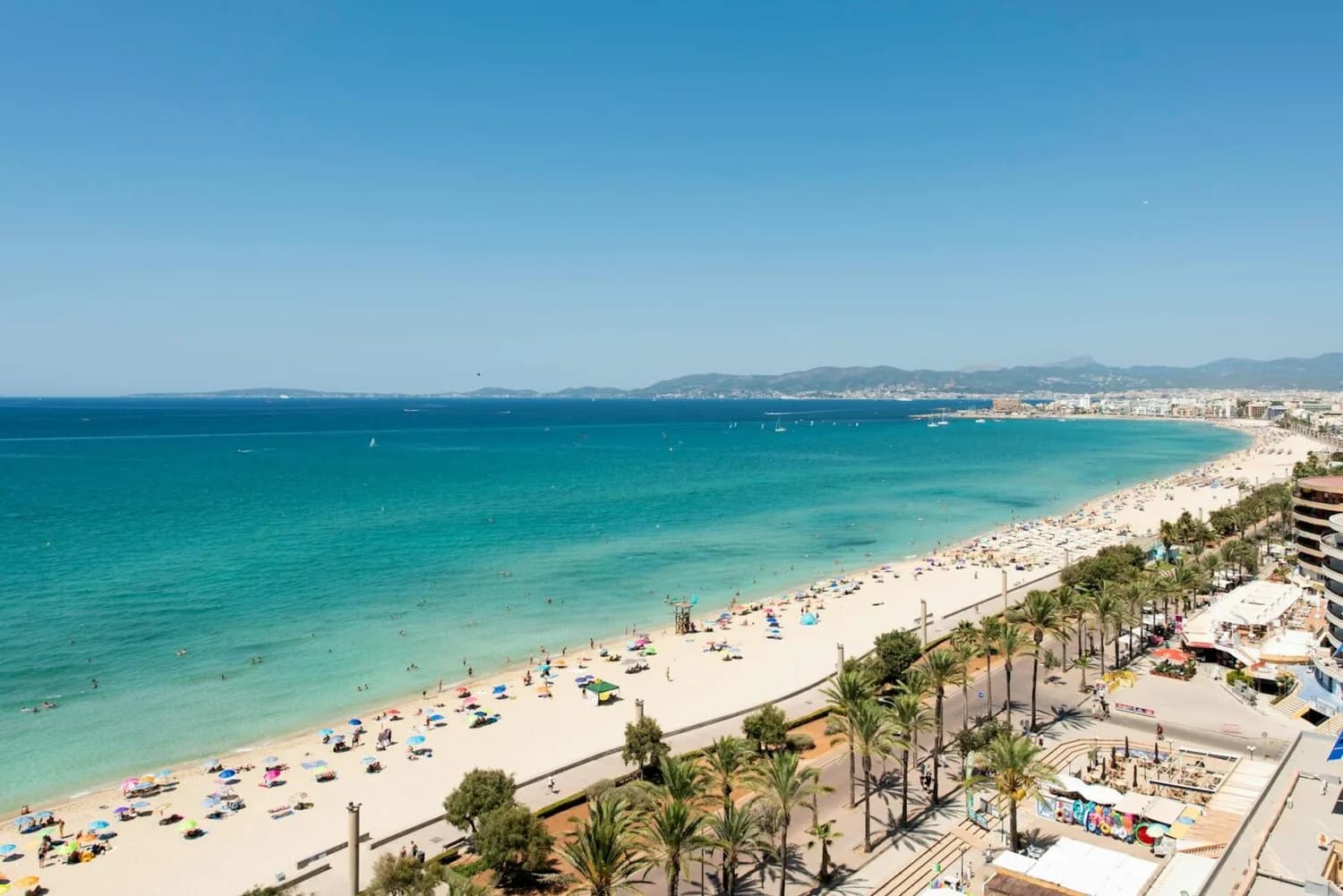Strandpromenade mit Palmen und Sonnenschirmen an einem langen Sandstrand auf Mallorca, türkisfarbenes Meer und entfernte Küstenlinie, wimmelnde Badegäste und Hotels nahe Flughafen Bremen-Reisenden Hinweis