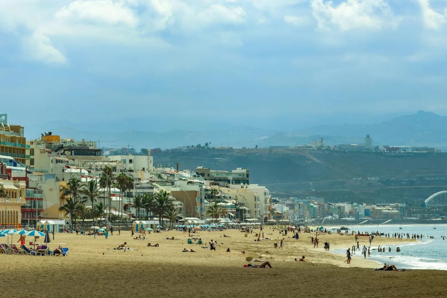 Strand von Las Palmas de Gran Canaria mit Sonnenschirmen, Badegästen, Promenade und Hochhäusern vor bergiger Küstenlandschaft