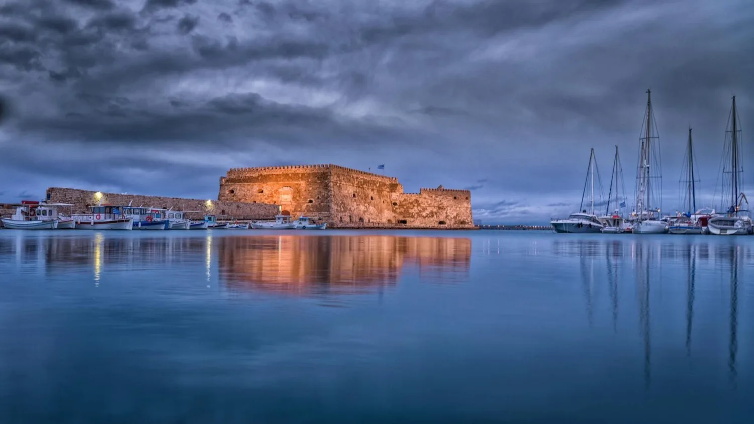 Hafen von Heraklion auf Kreta mit venezianischer Festung Koules, angelegten Fischerbooten und Segelyachten bei ruhigem, spiegelndem Wasser und bewölktem Himmel – Flugverbindung Nürnberg nach Heraklion.