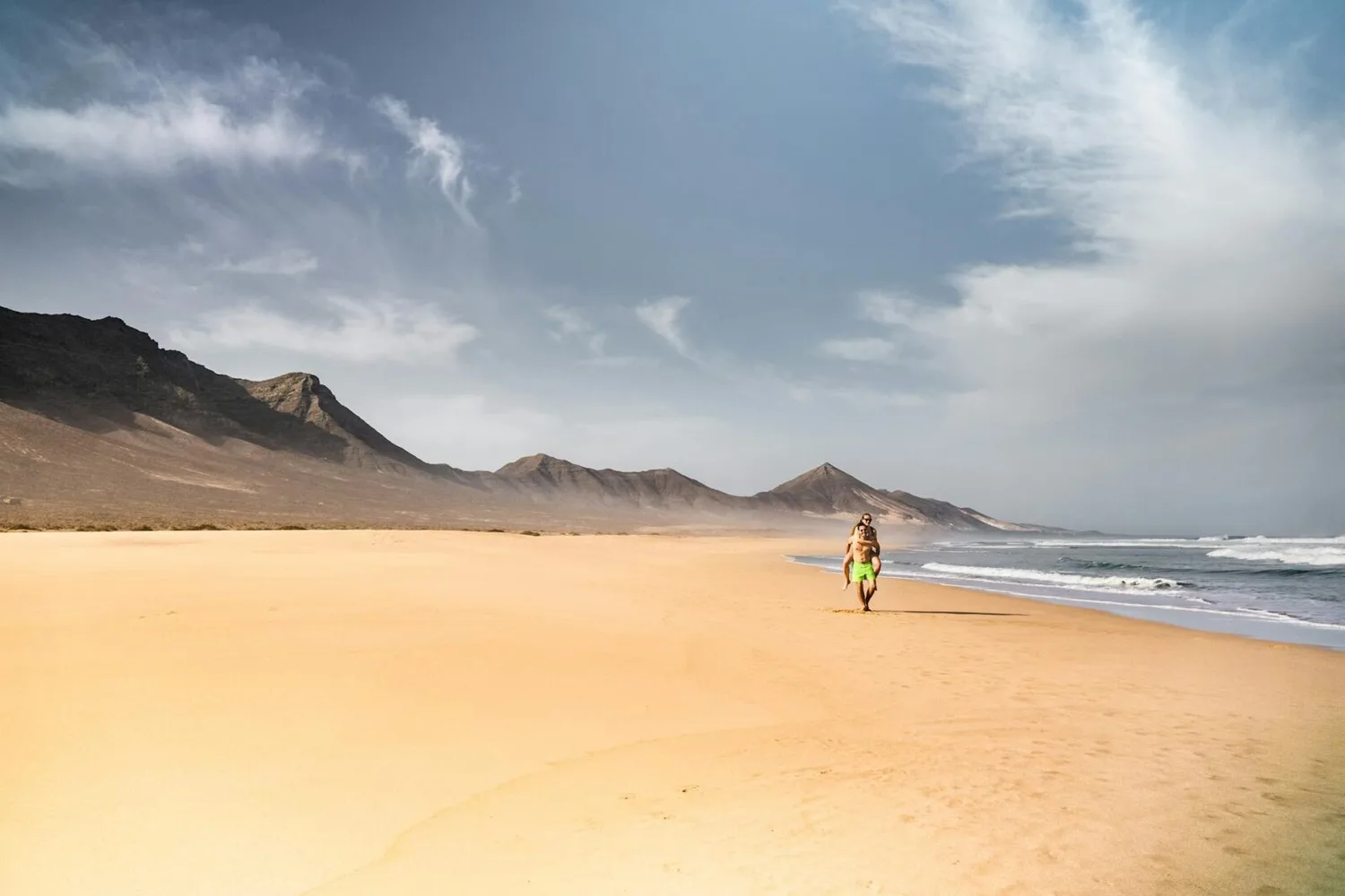 Paar läuft am einsamen Sandstrand in Spanien mit Bergen im Hintergrund und sanften Wellen am Ufer