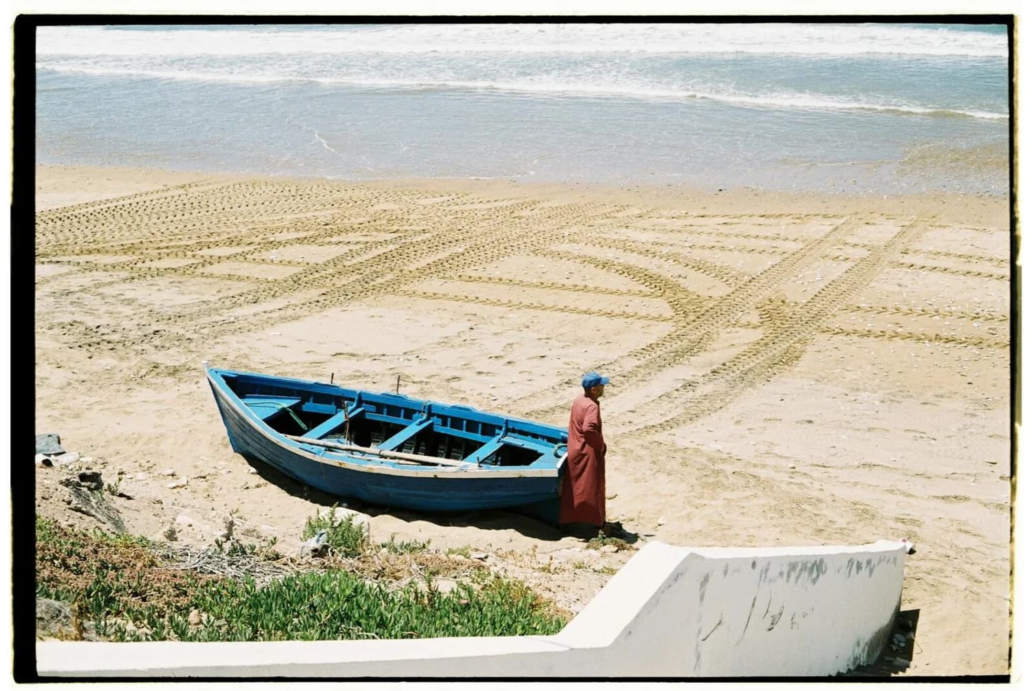 Fischer in roter Djellaba neben blauem Holzboot am Sandstrand von Tétouan, Marokko, mit Spuren von Reifenspuren im Sand und ruhiger Brandung im Hintergrund