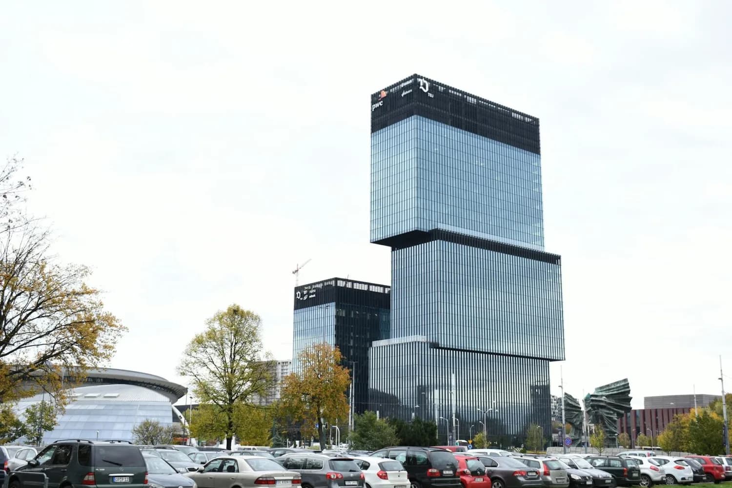 Modernes Bürohochhaus mit gestapelten Glasvolumen und Parkplatz im Vordergrund in Kattowitz, Bäume und umliegende Stadtsilhouette