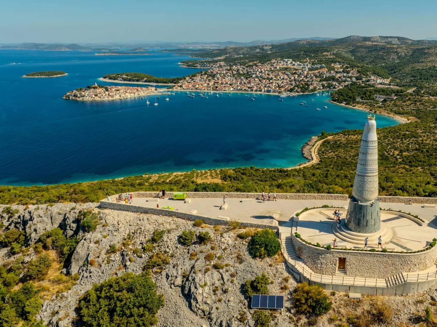 Aussicht auf kroatische Adriaküste mit blauem Meer, historischer Hafenstadt, Ankerboote und Aussichtspunkt mit großem Denkmal auf felsiger Klippe