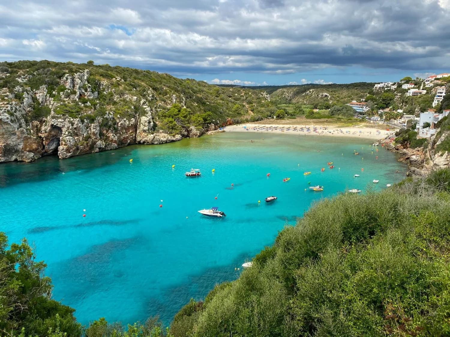Bucht mit türkisfarbenem Wasser, weißen Sandstrand, Booten und Kajaks, felsige Küste und Vegetation auf Menorca, Spanien